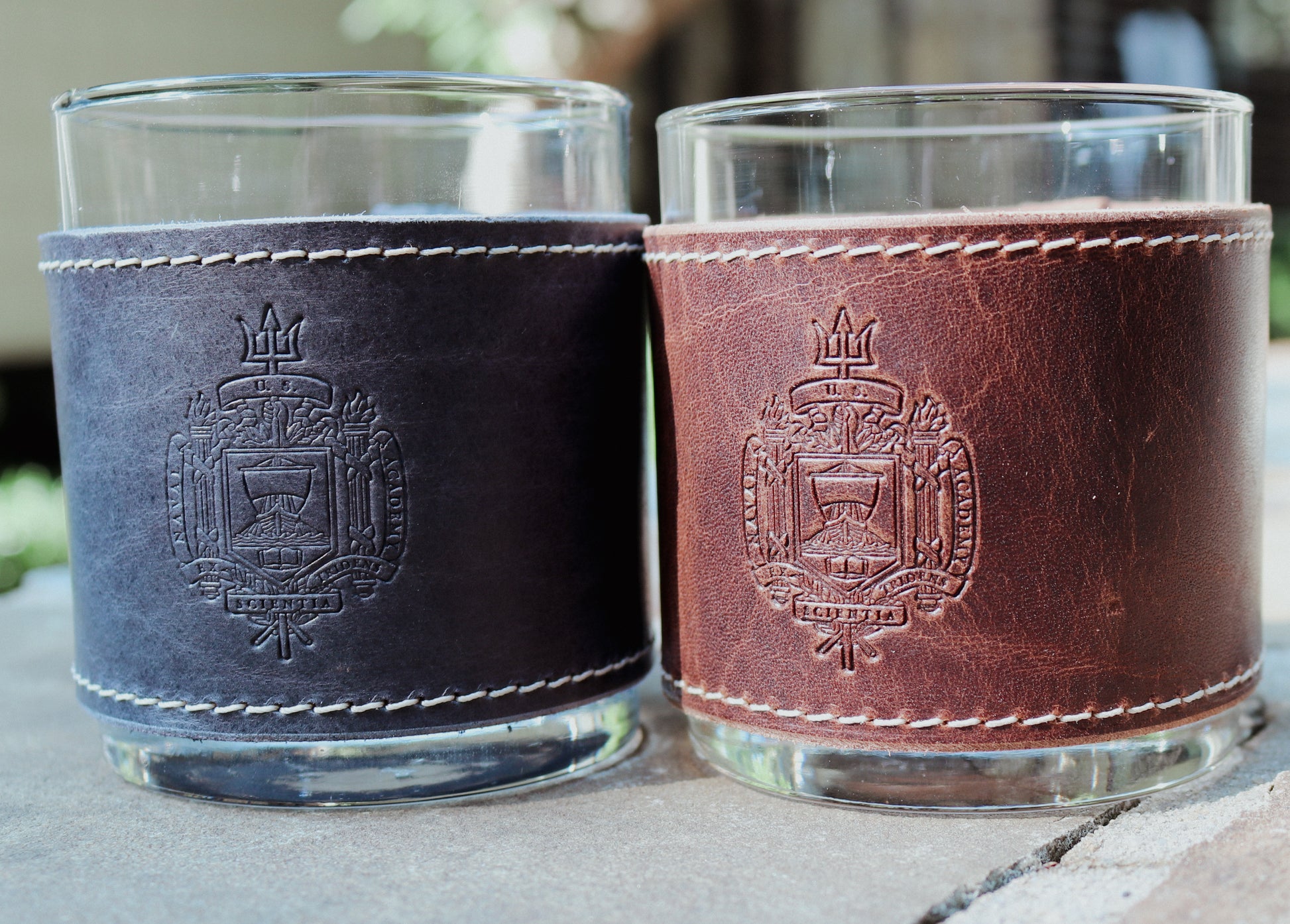 Two glass tumblers with leather sleeves featuring an embossed design on a stone surface.